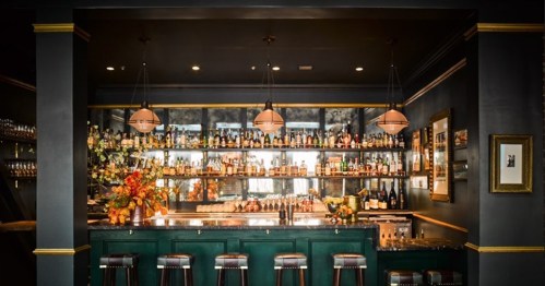 A stylish bar with a green counter, shelves of bottles, and warm lighting, featuring elegant decor and seating.