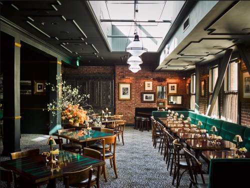 Elegant restaurant interior with wooden tables, green seating, and floral arrangements, featuring warm lighting and brick walls.
