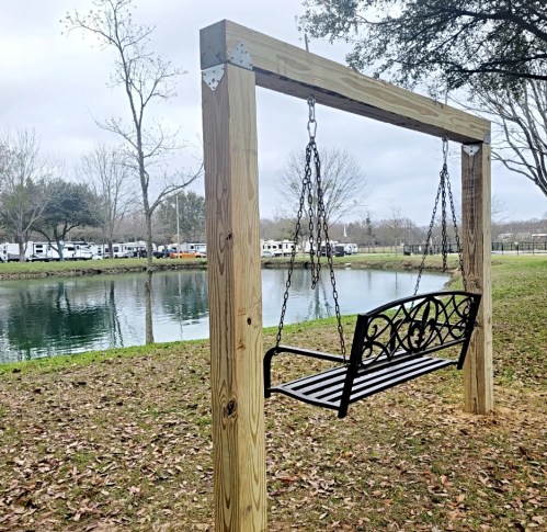 A wooden swing hangs from a frame by a pond, surrounded by trees and RVs in the background under a cloudy sky.