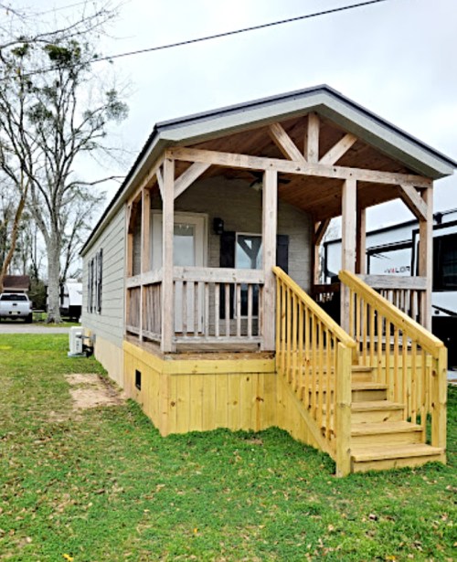 A small, modern cabin with a wooden porch and steps, surrounded by grass and trees on a cloudy day.
