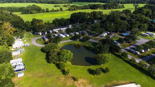 Aerial view of a campground with RVs, green fields, trees, and a small pond surrounded by grassy areas.