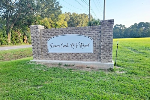 Sign for "Winners Circle RV Resort" on a brick structure, surrounded by green grass and trees.
