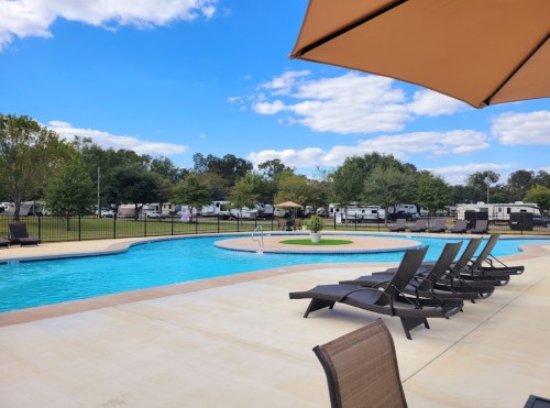 A sunny pool area with lounge chairs, surrounded by trees and RVs in the background under a blue sky with clouds.