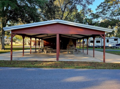 A covered picnic area with stone tables, surrounded by trees and RVs in a campground setting.