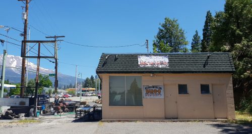 A small, light-colored building with a sign, surrounded by trees and mountains in the background on a sunny day.