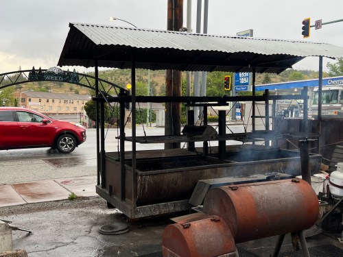 A roadside barbecue stand with smoke rising, next to a busy street and a red car passing by.