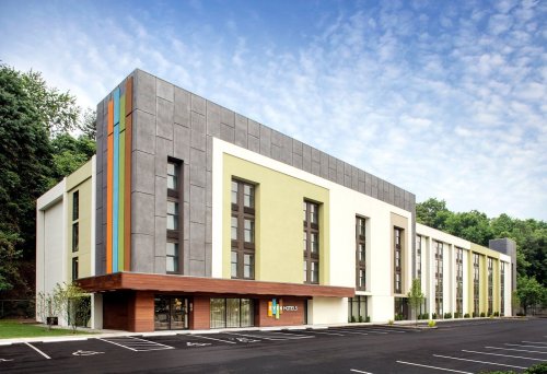 Modern hotel exterior with a mix of gray, green, and wood accents, surrounded by trees and a clear blue sky.