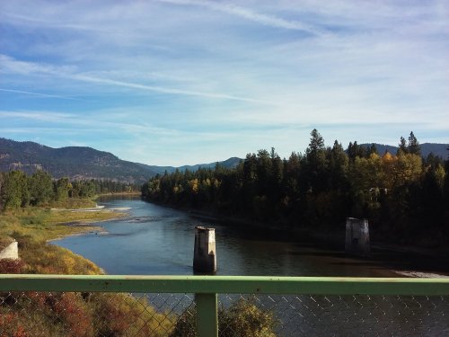 A serene river flows through a landscape of trees and mountains under a clear blue sky.