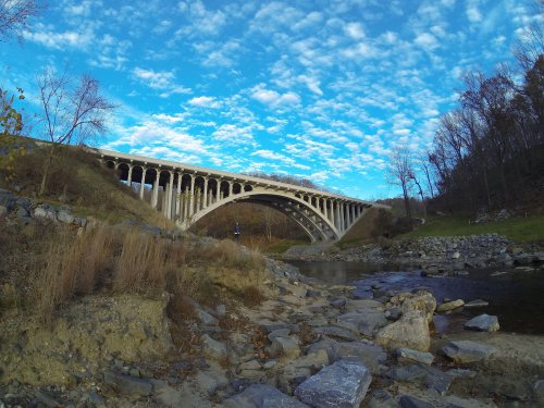A large concrete bridge arches over a river, surrounded by rocky banks and trees under a blue sky with scattered clouds.
