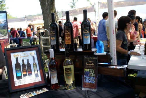 A display of wine bottles and a sign at a festival, with people mingling in the background.