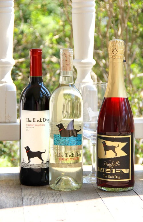 Three bottles of wine labeled "The Black Dog," featuring a red, a white, and a sparkling variety, displayed outdoors.