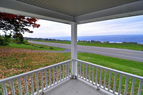 View from a porch overlooking a scenic landscape with a road, green fields, and distant hills under a cloudy sky.