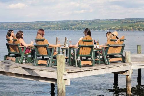 A group of women relaxes in Adirondack chairs on a dock by a lake, enjoying drinks with a scenic view.