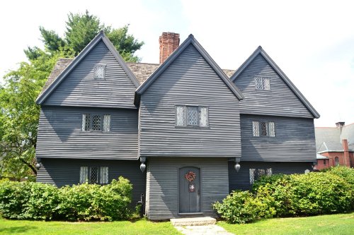 A dark gray, three-story house with a steep roof and decorative windows, surrounded by green shrubs and trees.