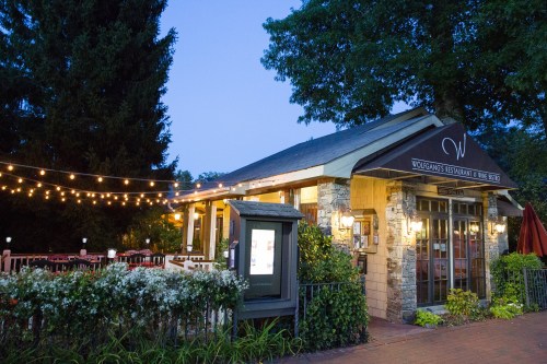 A cozy restaurant with outdoor seating, string lights, and a stone facade, surrounded by greenery at dusk.