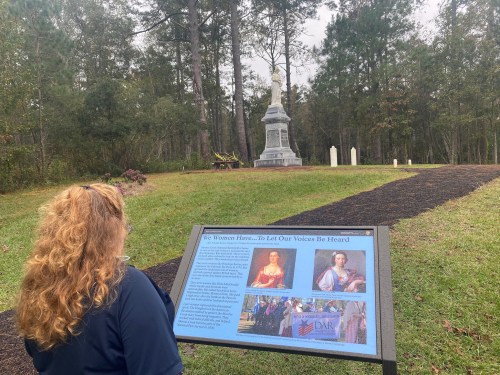 A woman reads a historical sign in front of a monument surrounded by trees and gravestones.