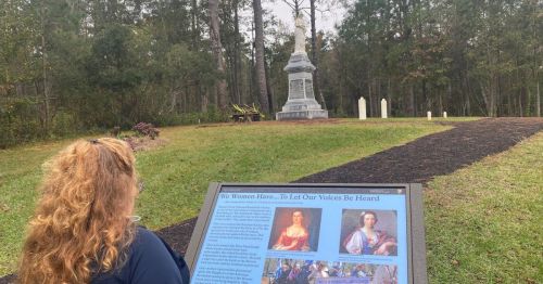 A woman reads a sign at a memorial site surrounded by trees, with a statue in the background.