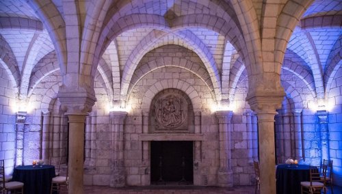 A stone-walled room with arched ceilings, softly lit in blue, featuring small tables and a decorative wall relief.