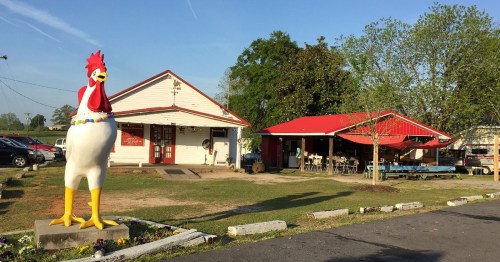 A large rooster statue stands in front of a rustic restaurant with a red roof and outdoor seating.