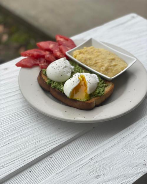 A plate with avocado toast topped with poached eggs, sliced tomatoes, and a small dish of creamy dip.