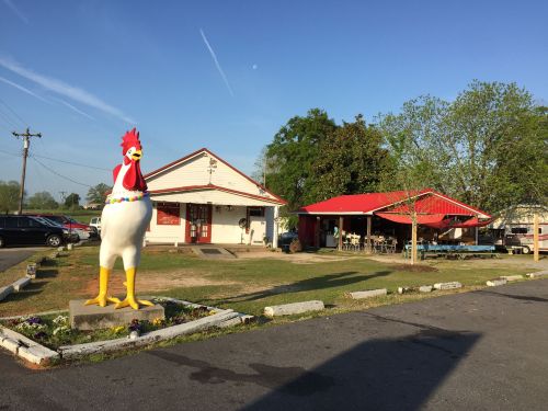 A large chicken statue stands in front of a restaurant with a red roof and outdoor seating area.