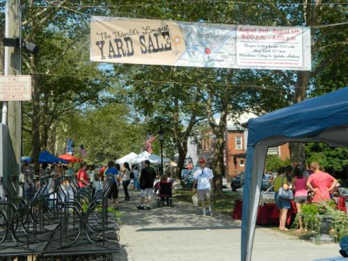 A bustling outdoor yard sale with vendors, shoppers, and a banner reading "The World's Largest Yard Sale."