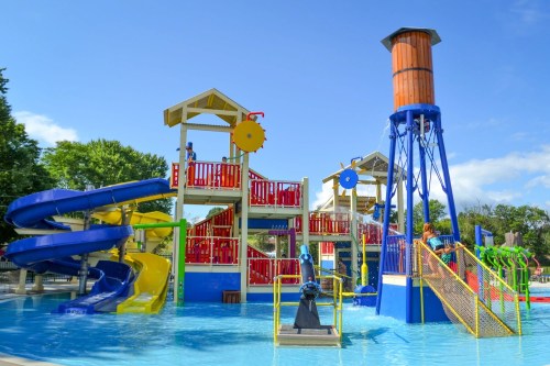 Colorful water park structure with slides, water features, and a large water tower, surrounded by a pool on a sunny day.