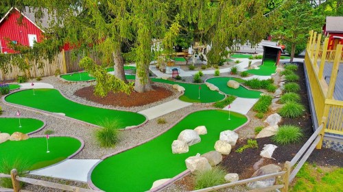 Aerial view of a mini-golf course with green turf, winding paths, rocks, and trees, surrounded by a rustic setting.