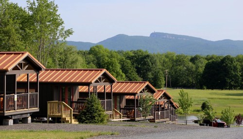 A row of cabins with metal roofs, set against a backdrop of green hills and a distant mountain.