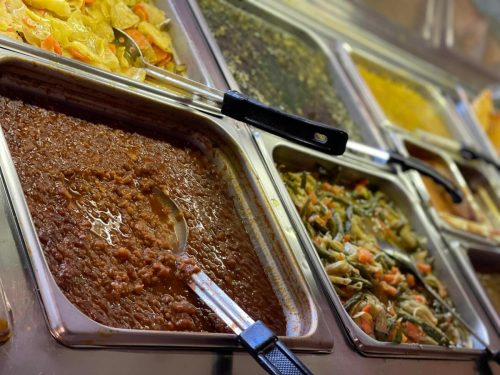 A variety of colorful dishes in metal trays, including lentils, vegetables, and sauces, with serving utensils.