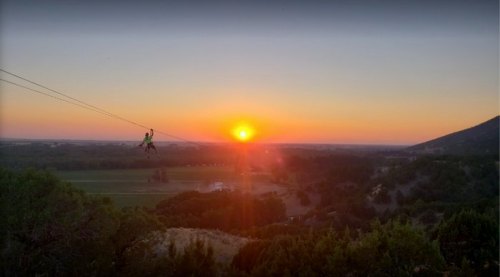 A person zip-lining against a vibrant sunset over a scenic landscape with hills and valleys.