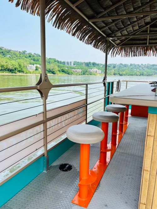 View of a riverside bar with stools, a thatched roof, and a scenic backdrop of greenery and water.