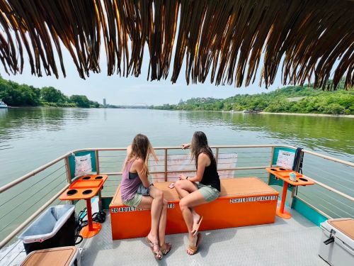 Two women sit on a boat, enjoying the view of a calm river surrounded by greenery under a thatched roof.