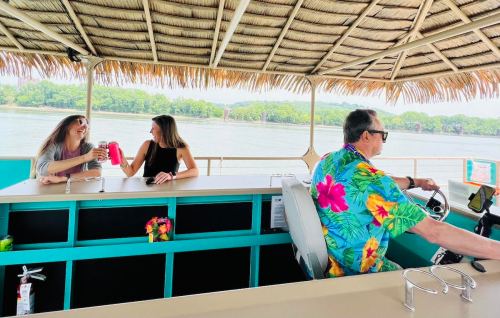 Two women enjoy drinks while chatting on a boat, with a man in a colorful shirt steering at the helm.