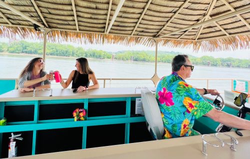 Two women enjoy drinks at a bar while a man steers a boat on a lake, surrounded by greenery and a thatched roof.