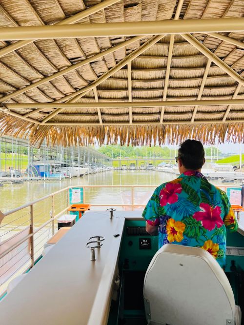 A person in a colorful floral shirt steers a boat under a thatched roof, with a marina in the background.