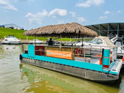 A tiki boat with a thatched roof docked at a marina, surrounded by boats and water, under a blue sky.