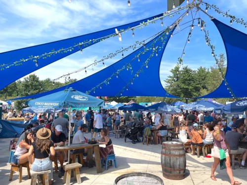 A lively outdoor gathering with people seated at tables under blue canopies, enjoying food and drinks in a festive atmosphere.