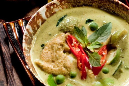 A bowl of green curry with chicken, green peas, red chili, and fresh basil leaves, garnished with lime.