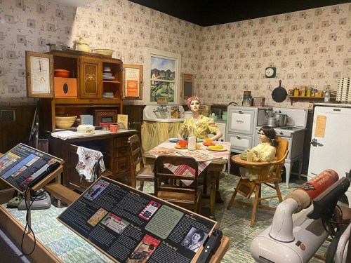 A vintage kitchen scene with a mannequin and child at a table, surrounded by retro appliances and decor.
