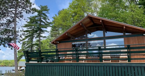 A wooden cabin with large windows, surrounded by trees, and an American flag waving in the breeze.
