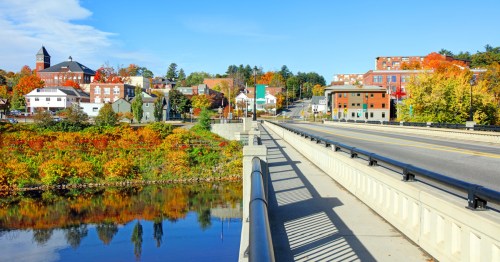 A scenic view of a town with colorful autumn foliage, a river, and buildings under a clear blue sky.