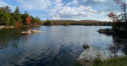A serene lake surrounded by trees with autumn foliage, under a partly cloudy sky. Rocks are visible in the foreground.