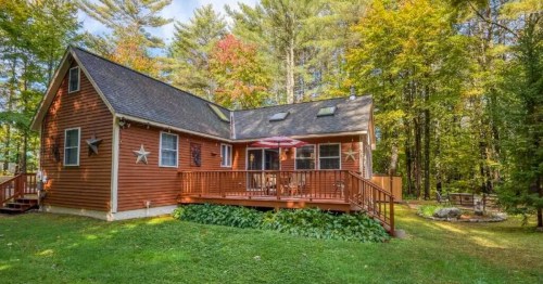 A cozy wooden house with a deck, surrounded by trees and greenery, featuring a patio umbrella and outdoor seating.