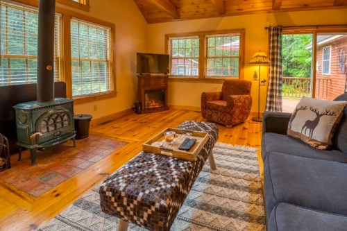 Cozy living room with a wood stove, comfortable seating, and large windows overlooking a forested area.