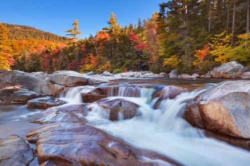 A serene river flows over smooth rocks, surrounded by vibrant autumn foliage and a clear blue sky.