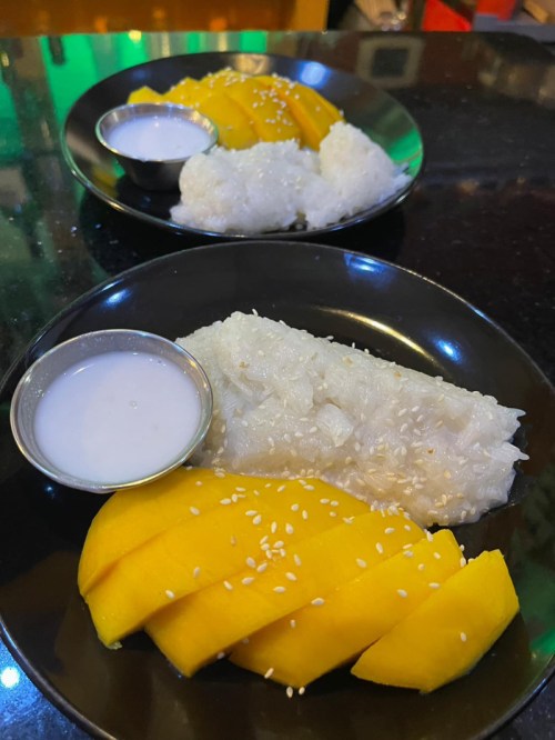 Two black plates with sticky rice, mango slices, and a small bowl of coconut milk, garnished with sesame seeds.