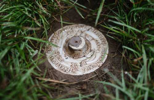 A white survey marker partially buried in grass and dirt, with engraved text and a metal bolt in the center.