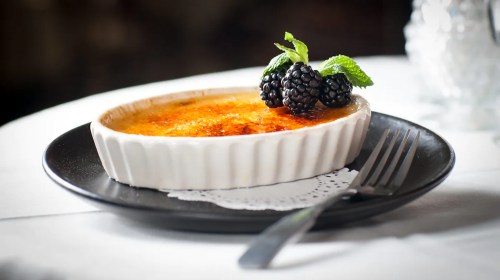 A creamy crème brûlée topped with blackberries and mint, served on a black plate with a fork beside it.