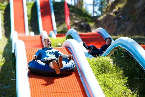 A person joyfully slides down a colorful water slide on an inflatable tube, surrounded by greenery and another slider in the background.